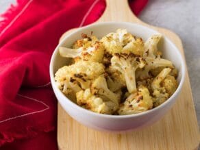 Frontal horizontal shot of roasted cauliflower in a dish resting on a small wood board with red fabric in background.