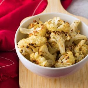 Frontal horizontal shot of roasted cauliflower in a dish resting on a small wood board with red fabric in background.