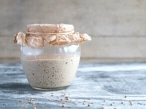 Glass jar of mature sourdough starter covered with crinkled paper and tied with twine, on a wooden countertop scattered lightly with rye seeds.