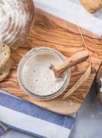 Overhead shot of sourdough starter with wooden spoon in glass bowl on a wooden cutting board. Sourdough boule sliced alongside the bowl. Dish of dry flour beside the cutting board. Scene is decorated by shaft of wheat.