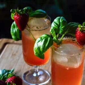 Two cocktail glasses filled with a red beverage and garnished with a sprig of basil and a strawberry.