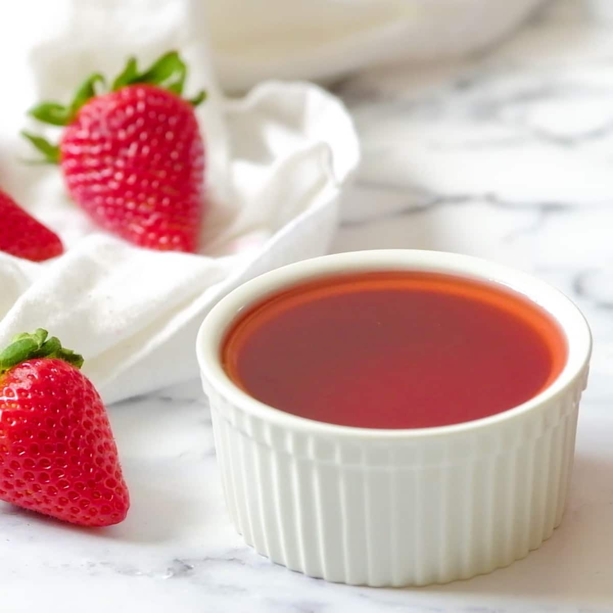 Square Crop - Strawberry Simple Syrup in a white dish on a white marble background with fresh strawberries.