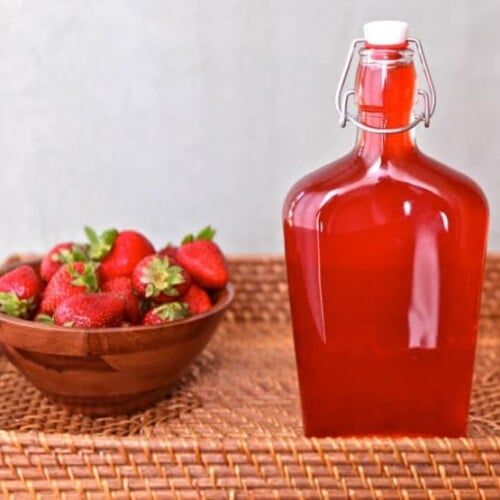 Strawberry jar next to a bowl of fresh strawberries inside a rectangular basket.