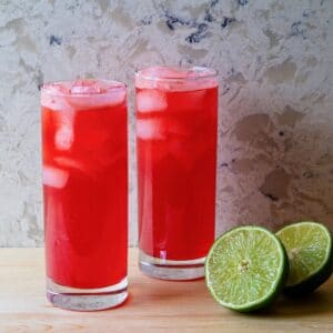 Horizontal shot of two red cocktails in tall glasses filled with ice. A sliced lime sits next to the glass on the right.