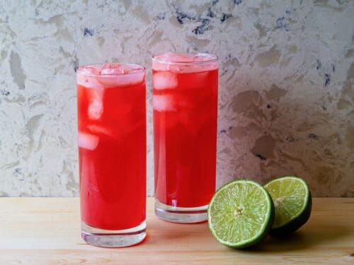 Horizontal shot of two red cocktails in tall glasses filled with ice. A sliced lime sits next to the glass on the right.