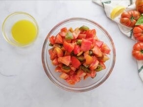 Glass bowl of chopped heirloom tomatoes with a small bowl of lemony olive oil dressing on the side, on a white marble background. Colorful heirloom tomatoes off to the right side, with a cloth napkin and lemon slices.