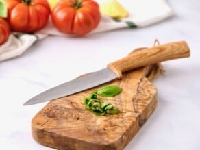 Chiffonade basil leaves on a wooden cutting board with a sharp knife with wooden handle. Heirloom tomatoes, lemon and linen towel in background.