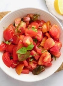 Overhead shot of a white bowl filled with fresh heirloom tomato salad topped with basil. Bowl rests on a wooden cutting board on a white marble background, with large fresh heirloom tomatoes, a linen towel, and a slice of lemon on the side.