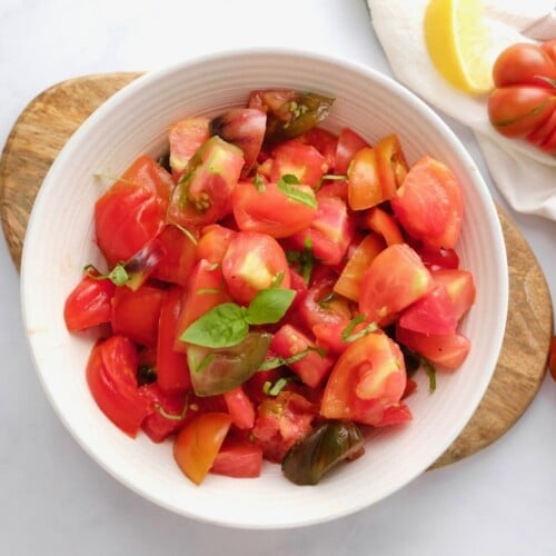 Overhead shot of a white bowl filled with fresh heirloom tomato salad topped with basil. Bowl rests on a wooden cutting board on a white marble background, with large fresh heirloom tomatoes, a linen towel, and a slice of lemon on the side.