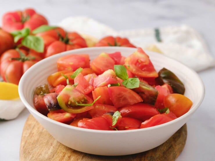 Horizontal close up shot of a summer heirloom tomato salad with fresh basil garnish in a white bowl on a wooden cutting board. Large ripe heirloom tomatoes and a cloth towel in background.