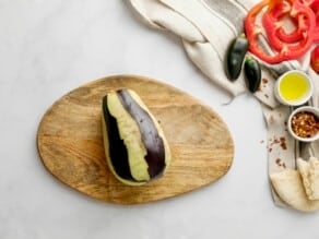 Medium eggplant with stem removed, peeled in strips so that part of the peel remains, while part of the peel is missing in long strips lengthwise down the eggplant. Linen napkin, small green peppers, red bell pepper slices, and chili flakes alongside the pot on the counter.