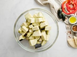 Cubed raw eggplant in a glass bowl. Linen napkin, small green peppers, red bell pepper slices, and chili flakes alongside the pot on the counter.