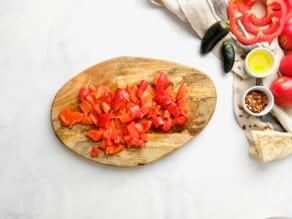 Roasted red pepper peeled and chopped on a wooden cutting board. Linen napkin, small green peppers, red bell pepper slices, and chili flakes alongside the pot on the counter.