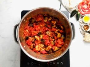 Pan of roasted eggplant salad cooked down and thickened. Cooking on a small portable burner on a marble countertop. Linen napkin, small green peppers, red bell pepper slices, and chili flakes alongside the pot on the counter.