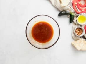 Small glass dish with paprika and olive oil mixed together. Linen napkin, small green peppers, red bell pepper slices, and chili flakes alongside the dish on the counter.