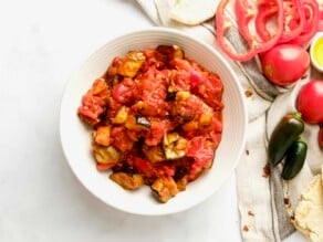 Overhead shot - a bowl of roasted eggplant salad with red peppers in a white bowl on a white marble countertop. Fresh tomatoes, dish of olive oil peppers and pita alongside the dish, with a linen napkin and scattered dried chili flakes.