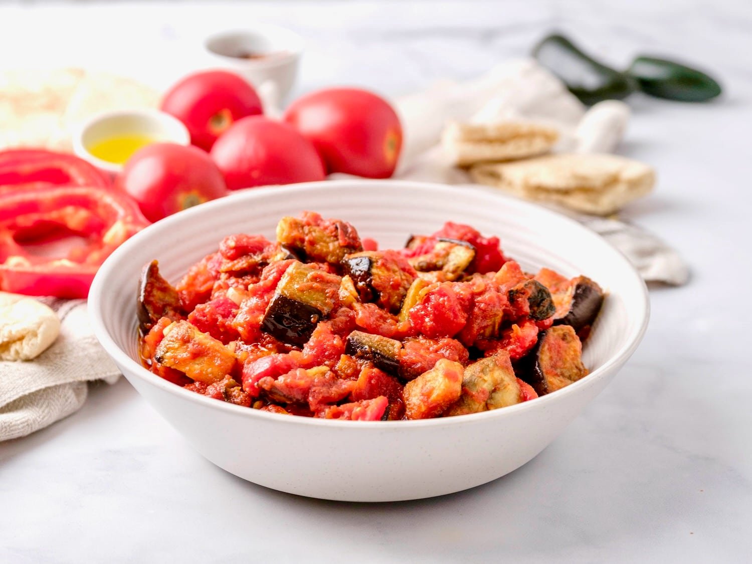 Featured wide horizontal shot - a bowl of roasted eggplant salad with red peppers in a white bowl on a white marble countertop. Fresh tomatoes, dish of olive oil peppers and pita in the background.