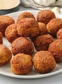 Horizontal image of a white plate filled with falafel balls. A bowl of tahini sauce, fresh herbs, a head of garlic, and pita bread sit in the background.