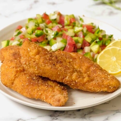 Close up - plate of chicken schnitzel, chopped salad in background with lemon, on marble counter.