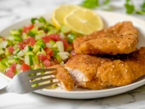 Close up - fork pulling a juicy piece from a crispy chicken schnitzel breast on a plate. Chopped Israeli salad in background with lemon and fresh parsley, on marble counter.