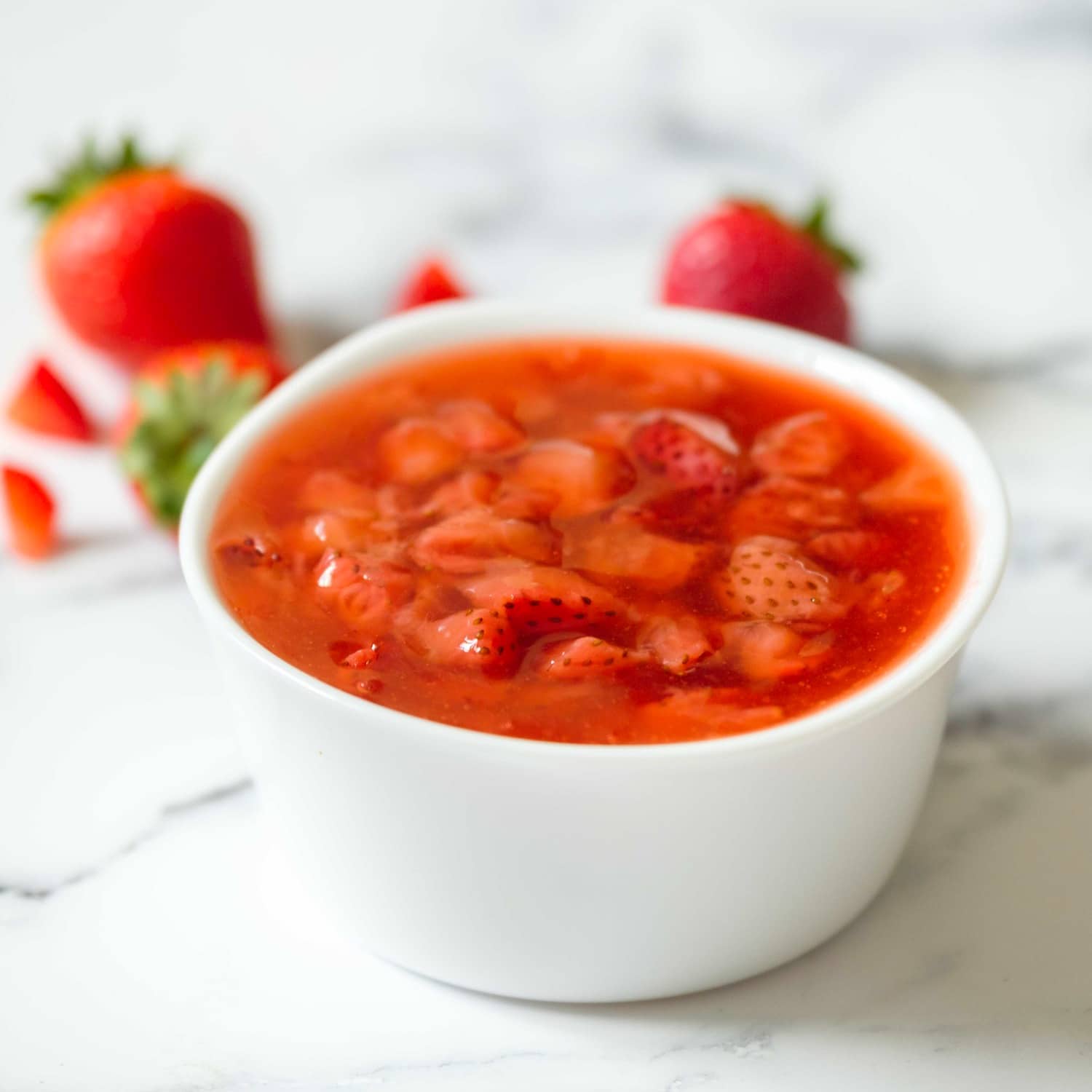 Square shot of a white dish of strawberry topping on a marble surface, strawberries in background.