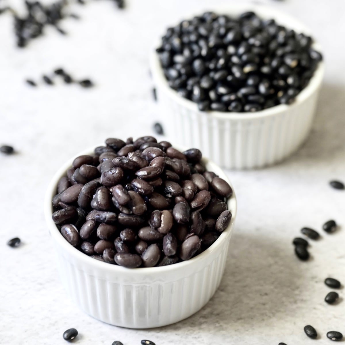 Square crop - two white dishes of cooked and uncooked black beans on marble background, dried black beans scattered behind.