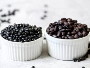 Two white dishes of cooked and uncooked black beans on marble background, dried black beans scattered behind.