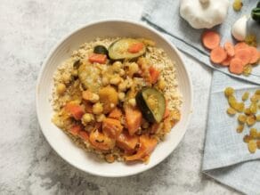 Overhead shot - dish of Moroccan Vegetable Couscous on marble surface with blue towel, raisins, apricots and garlic scattered beside it.