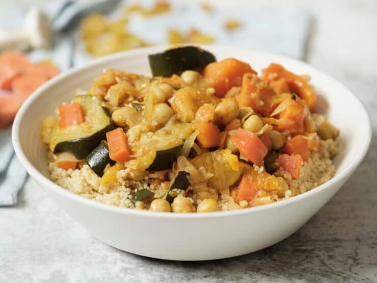 Close up horizontal shot - white dish of vegetable couscous with chickpeas, carrots, and zucchini on white marble background with towel, dry ingredients in backround - garlic, apricots, raisins.