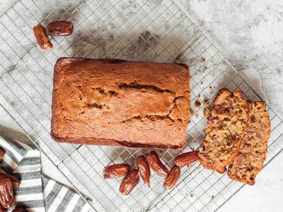 Wide horizontal shot - sliced date honey nut cake on cooling rack with luscious dates on marble countertop, striped linen towel laying beside the rack.