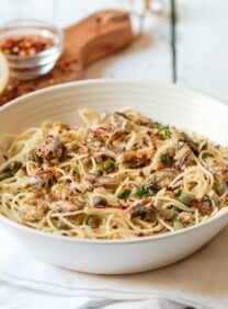 Square crop front view - white bowl of sardine pasta capellini garnished with chili flakes and fresh parsley, resting on a white cloth napkin, wooden cutting board in background with lemon half and chili flakes, on white wood table.