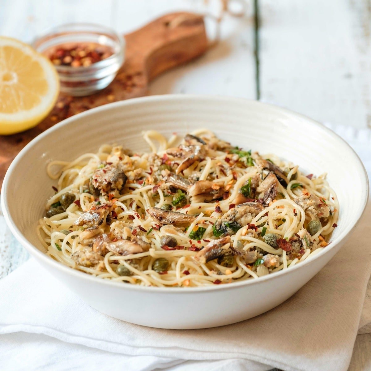 Square crop front view - white bowl of sardine pasta capellini garnished with chili flakes and fresh parsley, resting on a white cloth napkin, wooden cutting board in background with lemon half and chili flakes, on white wood table.