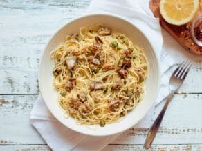 Overhead view - dish of Mediterranean sardine pasta with chili flakes, olive oil, lemon and capers on white linen napkin on distressed wood table, lemon half and dish of chili flakes on the side on a wooden cutting board.