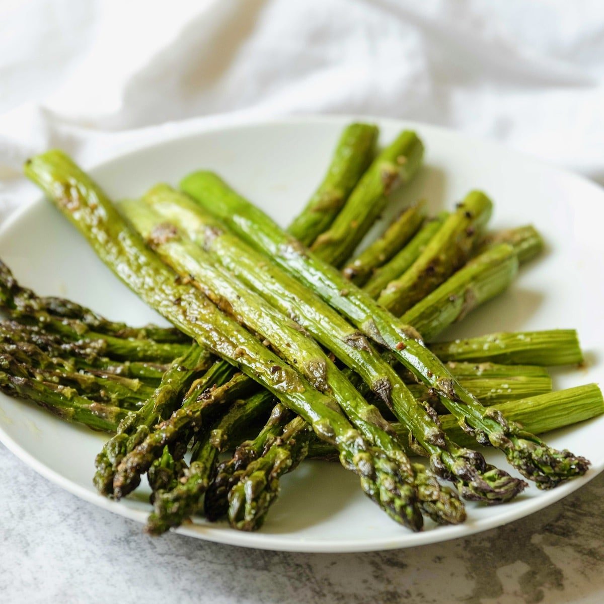 Square Crop - Three small piles of aligned roasted asparagus criss-crossed diagonally on a white plate, linen napkin in background.