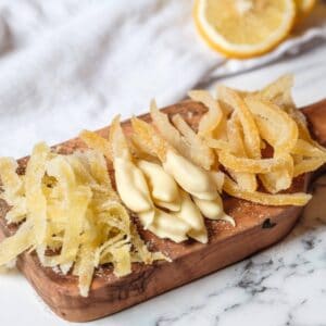 Horizontal shot - wide shot of candied lemon peels on a small wood cutting board on marble counter. Large peels, small peels, peels dipped in white chocolate. Lemon half and white linen towel in background.