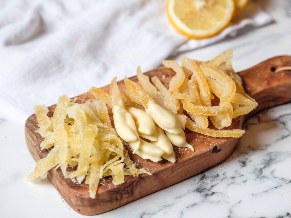 Horizontal shot - wide shot of candied lemon peels on a small wood cutting board on marble counter. Large peels, small peels, peels dipped in white chocolate. Lemon half and white linen towel in background.