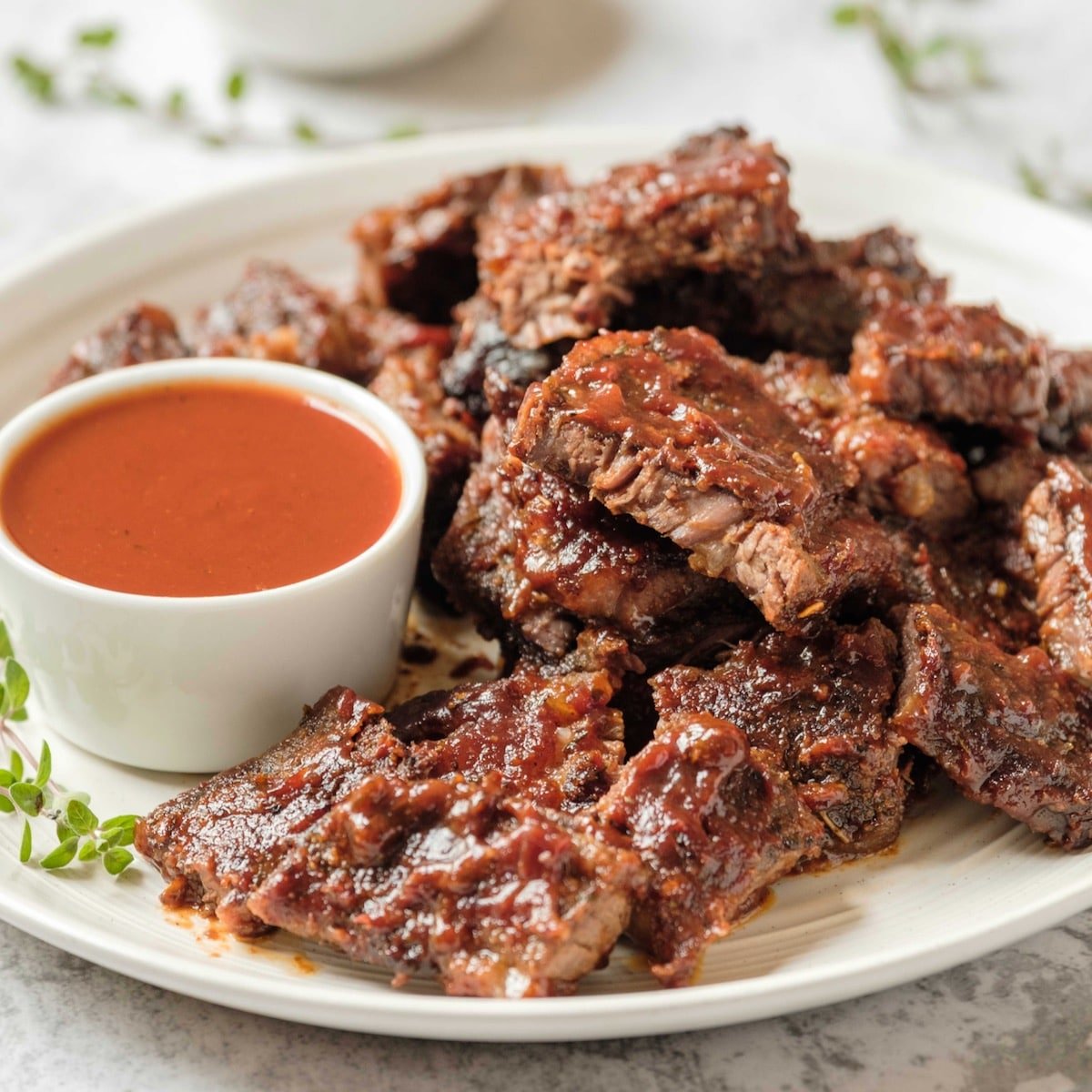 Square Crop- White plate of oven barbecued short ribs with sauce, garnished with fresh thyme, on a marble countertop.