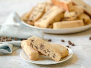 Small plate of chocolate chip mandel bread aka mandelbrot cookies - three piled in front of a larger plate of cookies in background, as well as a towel and chocolate chips in background.