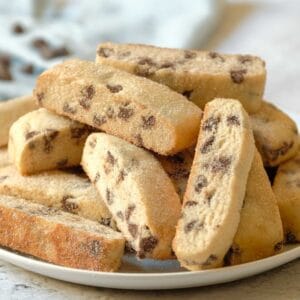 Horizontal shot - plate piled high with Jewish mandel bread cookies, aka mandelbrot, with towel and chocolate chips in background, on a marble countertop.