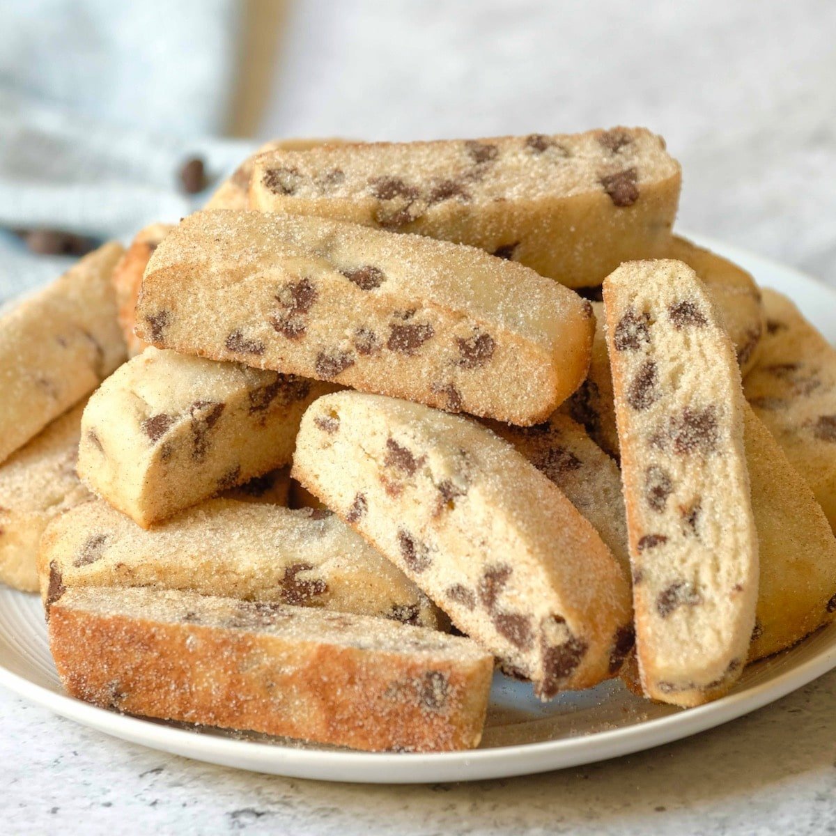 Square featured shot - plate piled high with Jewish mandel bread cookies, aka mandelbrot, with towel and chocolate chips in background, on a marble countertop.