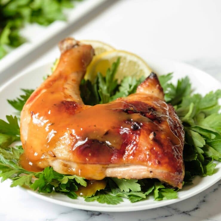 Square Featured Shot - whole roast chicken leg topped with honey garlic sauce on a bed of parsley garnish on white plate, platter of chicken in background, on a marble countertop.