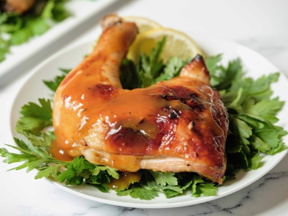 Horizontal shot - whole roast chicken leg topped with honey garlic sauce on a bed of parsley garnish on white plate, platter of chicken in background, on a marble countertop.