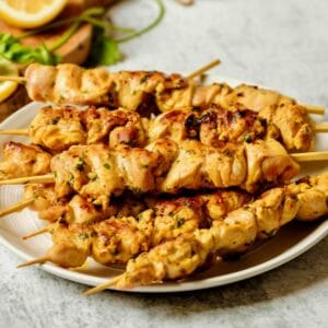 Horizontal shot - plate of Lemony Marinated Chicken Skewers on a marble countertop, wood cutting board with lemon and parsley in background.