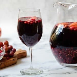 Horizontal shot - Glass of dark red Rosh Hashanah Sangria with a pitcher of sangria in background, each with fruit pieces floating on top. Wood cutting board with grapes and pomegranate arils, half a pomegranate in background.