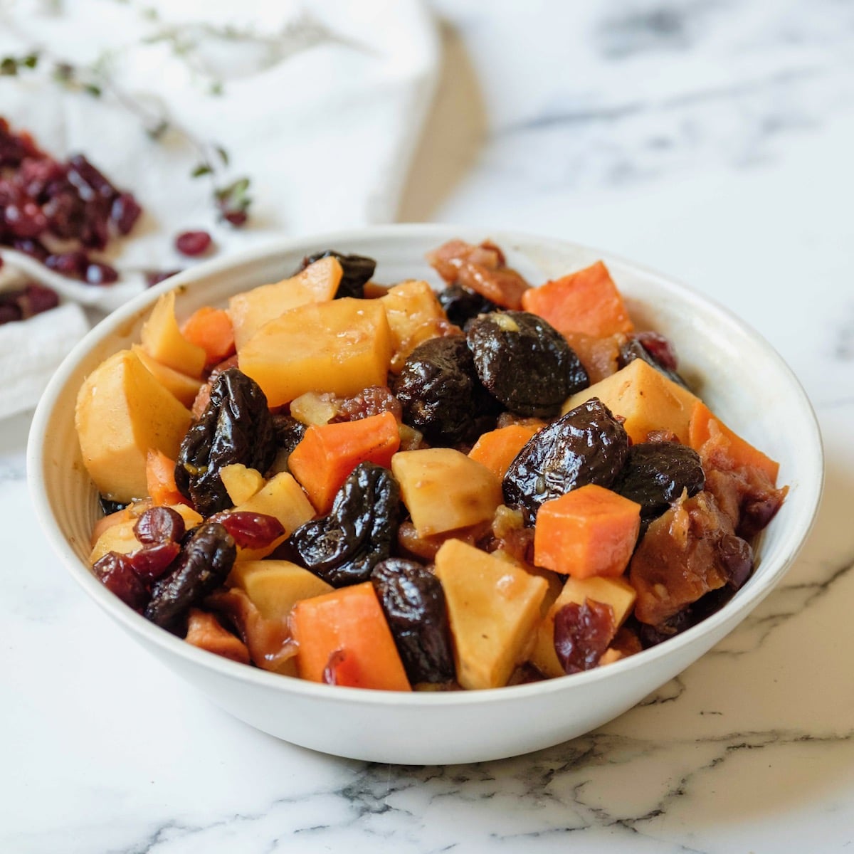 Square featured shot - white bowl of tzimmes with sweet potatoes, prunes, carrots, and dried cranberries on a marble countertop, linen napkin with dried cranberries in background.