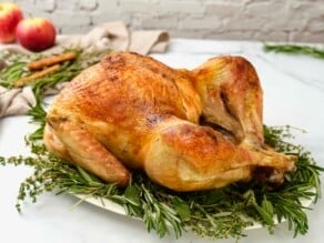 A platter of a whole classic roast turkey browned to perfection, on a bed of fresh green aromatic herbs. Apples, herbs, cinnamon sticks, and linen napkins in background.