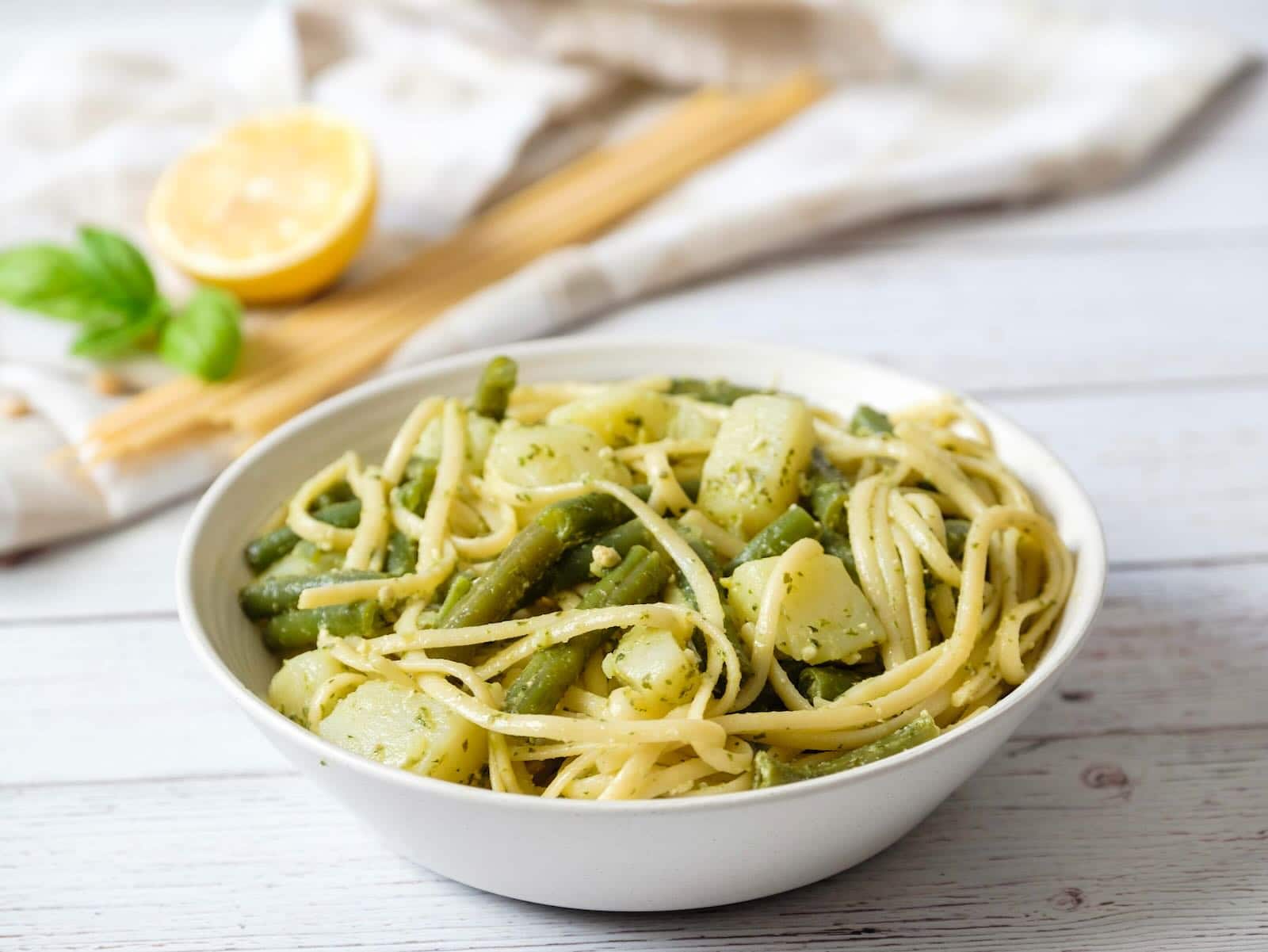 Vertical wide shot of trenette al pesto - delicious pasta recipe with trenette or linguine pasta, potatoes, and green beans in a white bowl on a marble counter. Lemon and basil with towel in background.