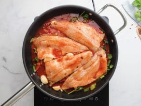 Horizontal overhead shot of a black sautee pan containing four white fish fillets laying on top of a bed of greens, red peppers, and garlic cloves.