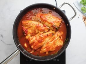 Horizontal overhead shot of a black sautee pan containing four white fish fillets laying on top of a bed of greens, red peppers, and garlic cloves.