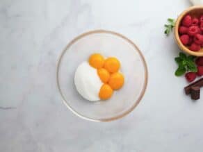 Overhead horizontal shot of a glass mixing bowl containing egg yolks and white sugar.